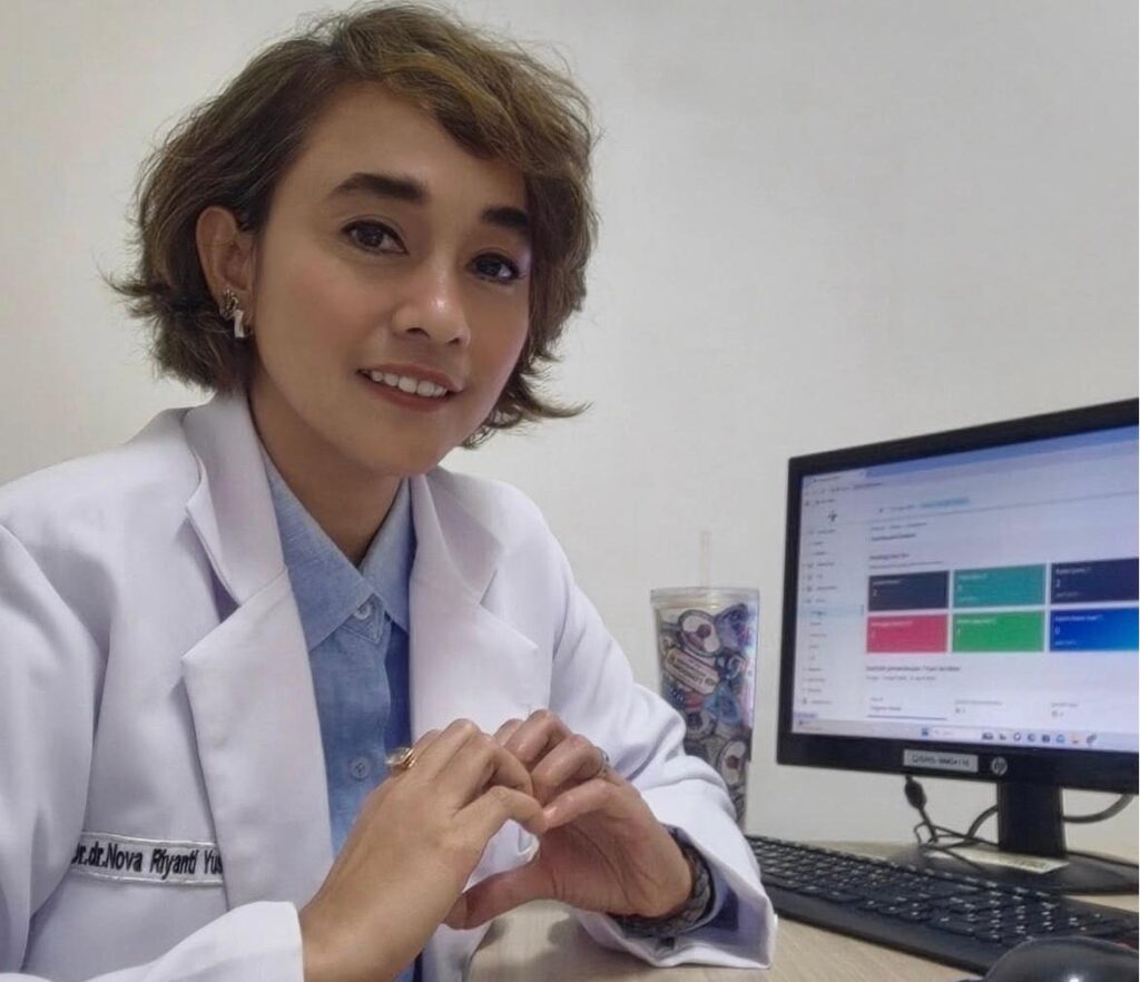 Dr Nova riyanti yusuf spkj catrawarta Female doctor in a white coat sits at a desk smiling at the camera with a computer monitor showing colorful tiles nearby